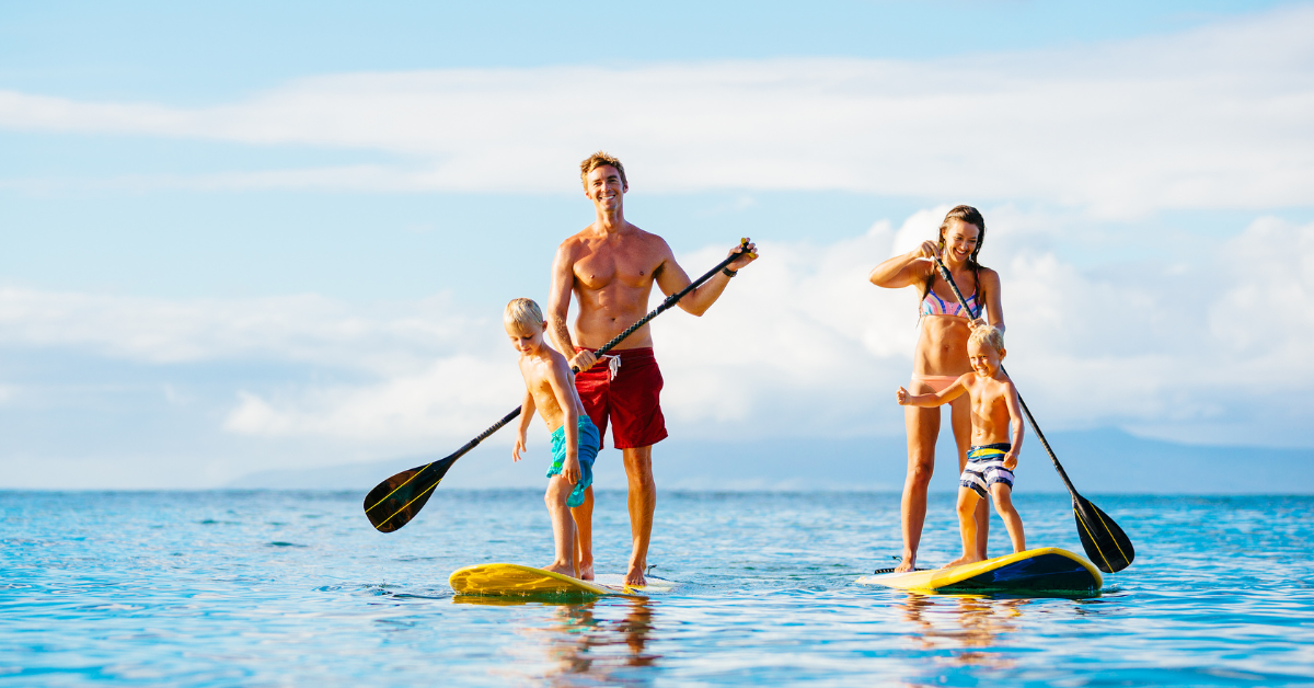 A smiling family enjoys paddleboarding together on calm bay waters near Brigantine, NJ. The clear blue sky and distant shoreline set the perfect backdrop for a fun coastal vacation—ideal for promoting Brigantine vacation rentals on ShoreSummerRentals.com.